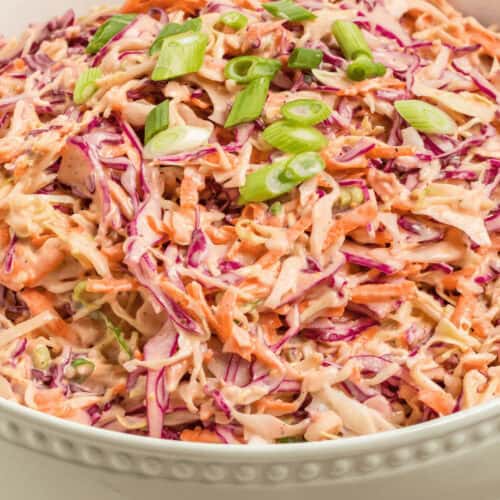 A bowl of spicy coleslaw made with shredded purple and white cabbage, carrots, and a creamy dressing, topped with sliced green onions.