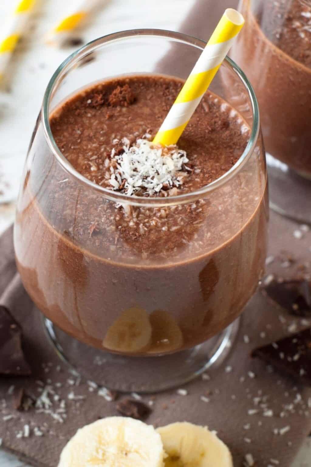 A glass of Chocolate Peanut Butter Banana Smoothie with a yellow-and-white striped straw, topped with shredded coconut and surrounded by banana slices and chocolate pieces.