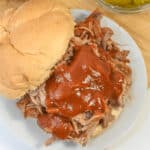 Dr. Pepper Pulled Pork sandwich topped with barbecue sauce on a bun, served on a white plate with a side of sliced pickles in a bowl.
