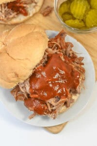 Dr. Pepper Pulled Pork sandwich topped with barbecue sauce on a bun, served on a white plate with a side of sliced pickles in a bowl.