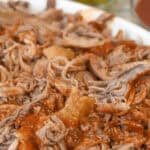 Close-up on the meat with barbecue sauce on a white plate, accompanied by a small bowl of pickles in the background.