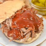 A sandwich with barbecue sauce on a white plate next to a small bowl of pickles.