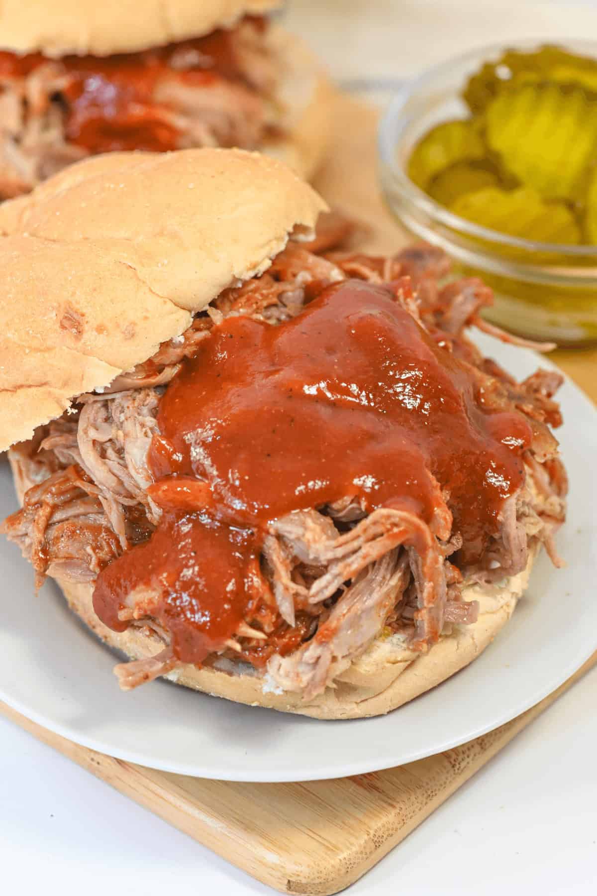 A sandwich with barbecue sauce on a white plate next to a small bowl of pickles.