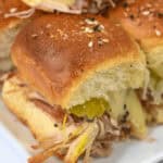 Close-up of a plate with several Cuban sliders, topped with pickles and melted cheese, served on soft buns.