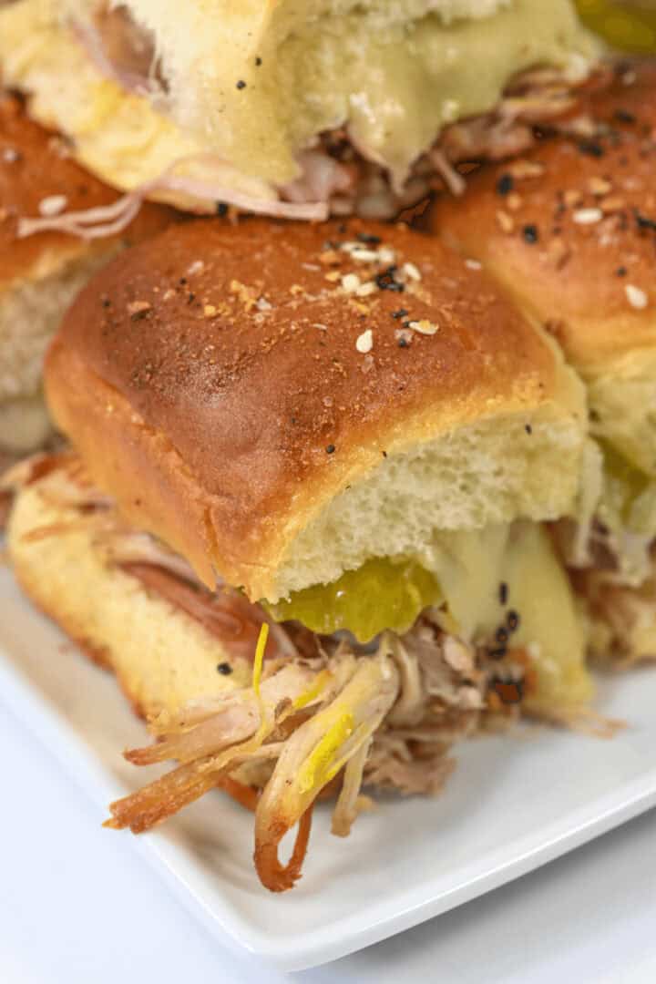 Close-up of a plate with several Cuban sliders, topped with pickles and melted cheese, served on soft buns.