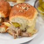 A close-up of the sandwiches with shredded meat, melted cheese, and pickles on a soft bun, topped with seasonings, all served on a white plate.