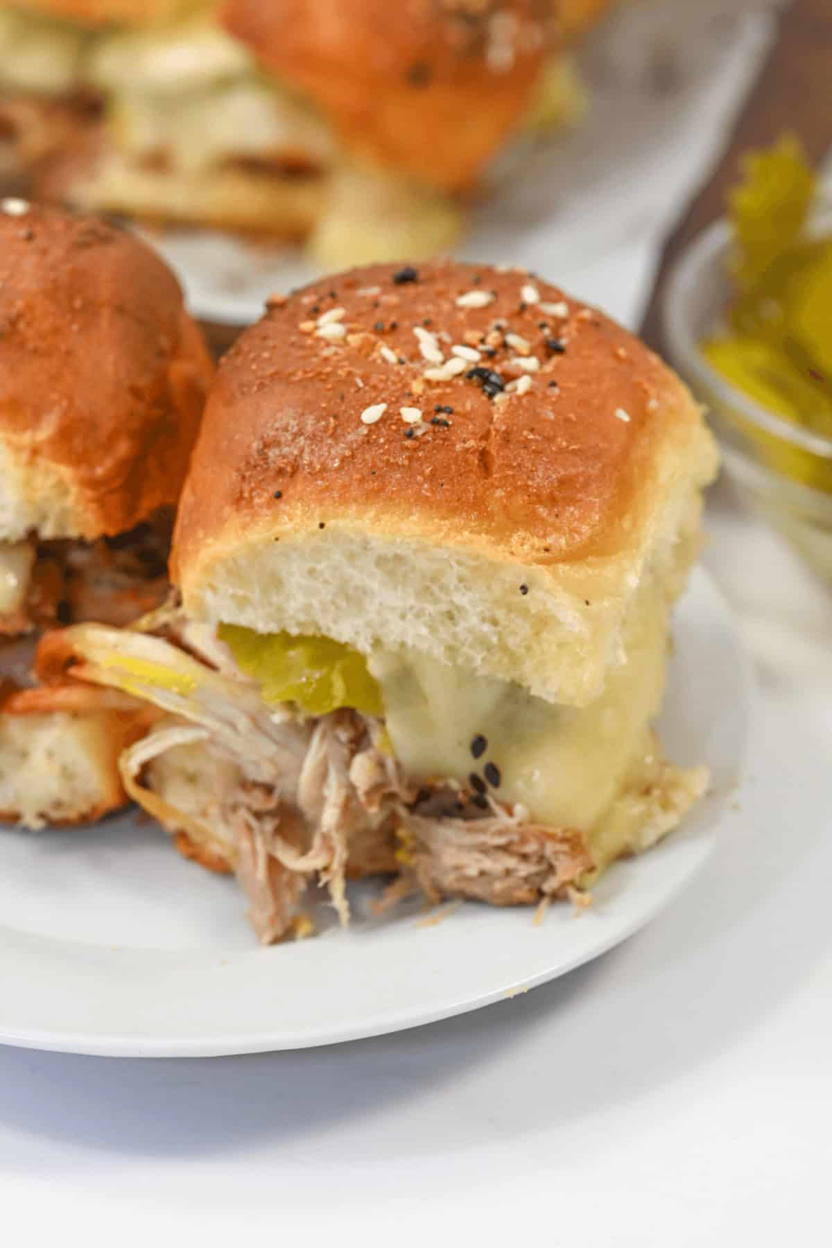 A close-up of the sandwiches with shredded meat, melted cheese, and pickles on a soft bun, topped with seasonings, all served on a white plate.