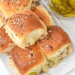A plate of food made with seeded bread rolls, filled with pulled pork and melted cheese. A small bowl of pickles is placed beside the sandwiches.