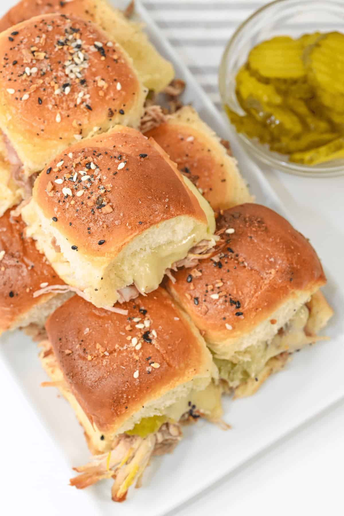 A plate of food made with seeded bread rolls, filled with pulled pork and melted cheese. A small bowl of pickles is placed beside the sandwiches.