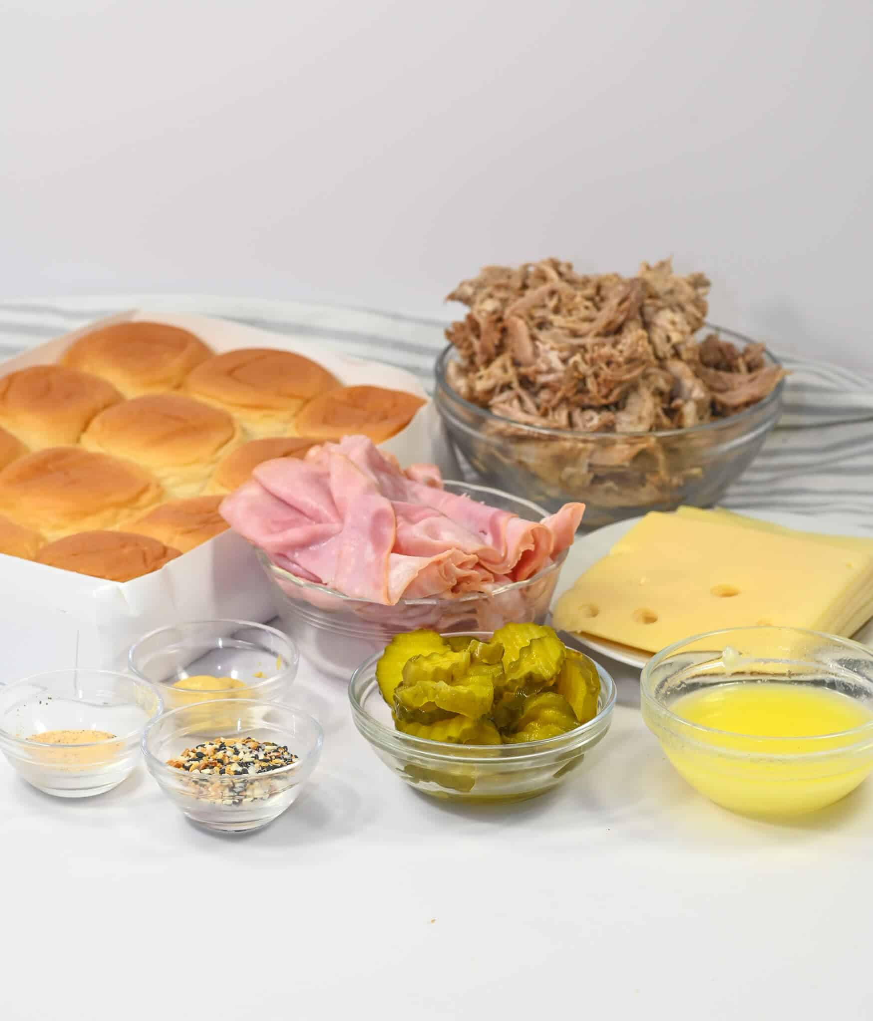 Ingredients for Cuban Sliders include slider rolls, sliced ham, shredded pork, Swiss cheese, pickles, and bowls of butter, seasoning, and spices laid out on a countertop.