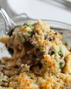 A close-up of an old-fashioned tuna casserole dish with a fork, featuring layers of ingredients including peas and mushrooms, topped with a toasted breadcrumb crust.