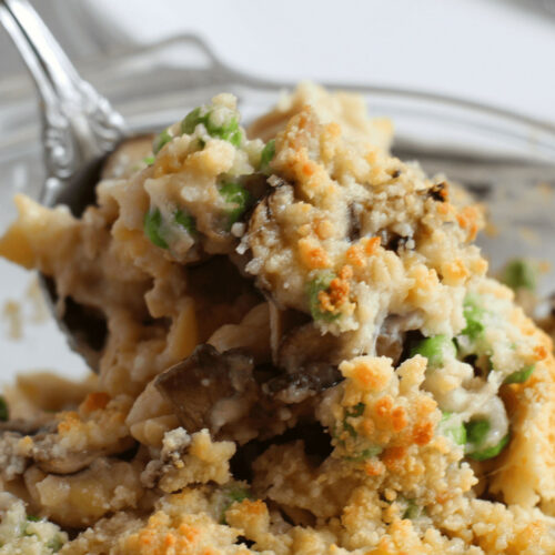 A close-up of an old-fashioned tuna casserole dish with a fork, featuring layers of ingredients including peas and mushrooms, topped with a toasted breadcrumb crust.
