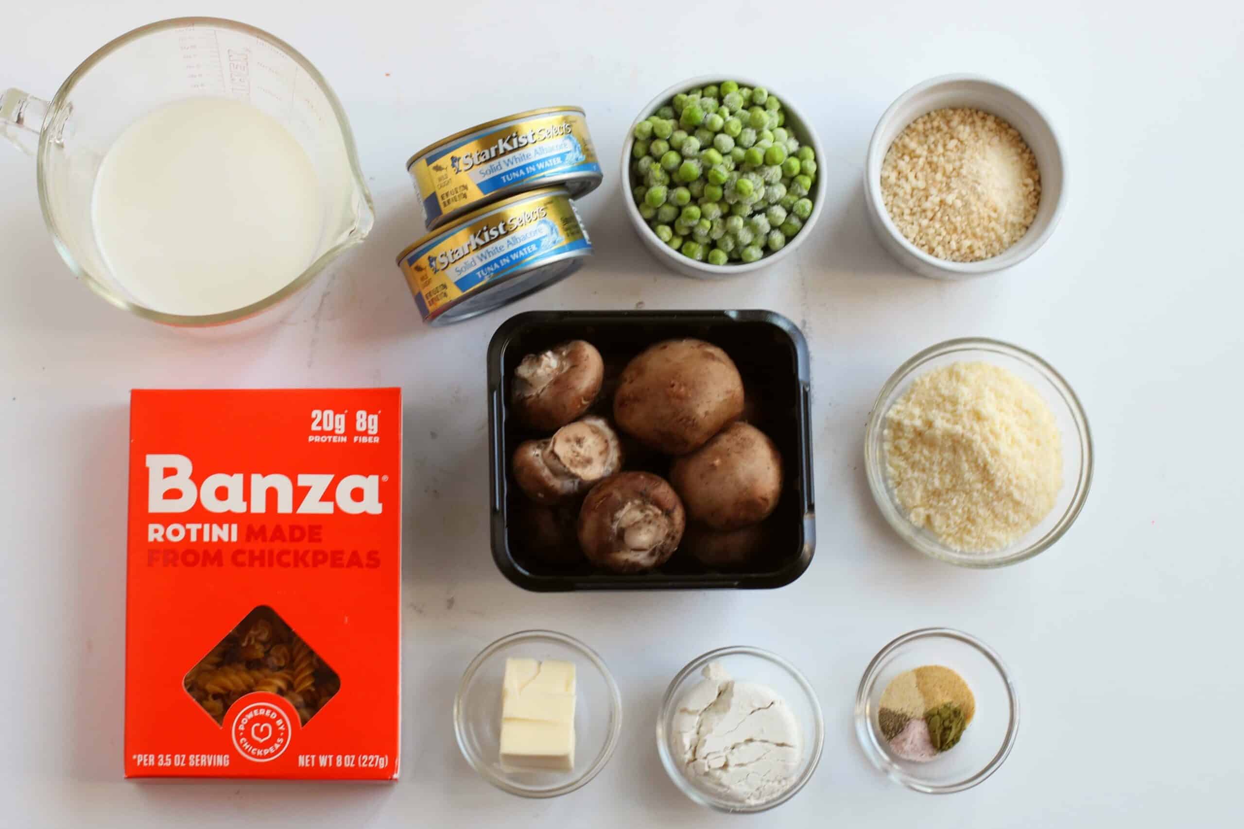 Ingredients for an old-fashioned tuna casserole recipe laid out on a white surface, including chickpea pasta, mushrooms, peas, cheese, and various seasonings.