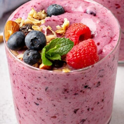 A glass of thick Berry Banana Smoothie topped with blueberries, raspberries, granola, almonds, and a sprig of mint.