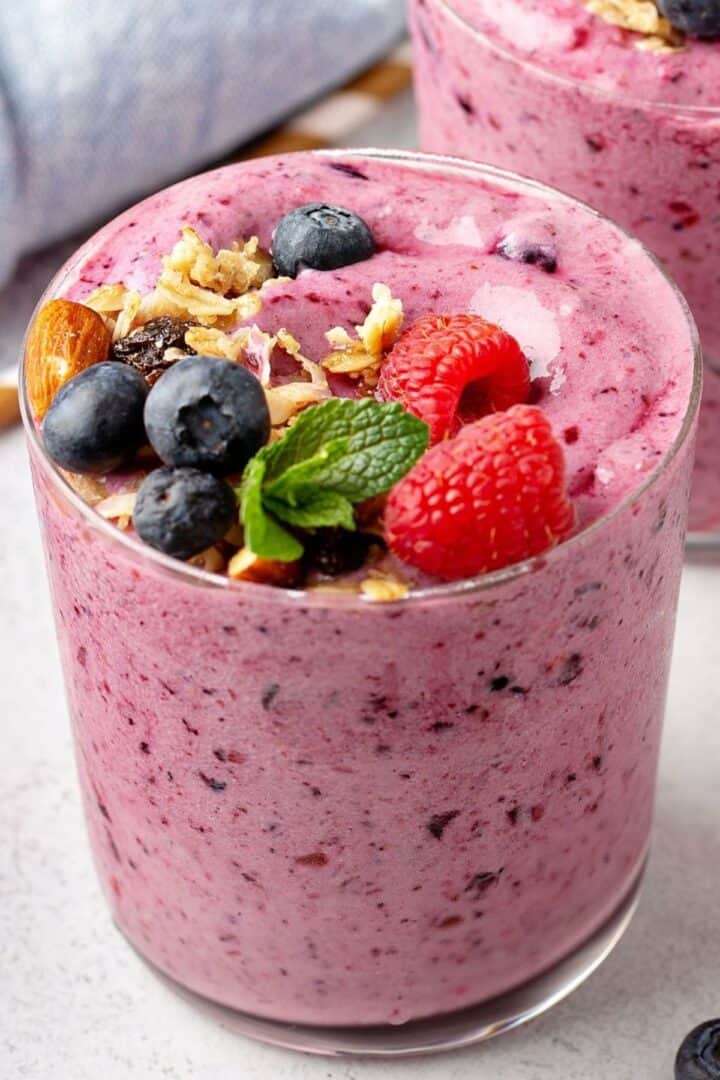A glass of thick Berry Banana Smoothie topped with blueberries, raspberries, granola, almonds, and a sprig of mint.