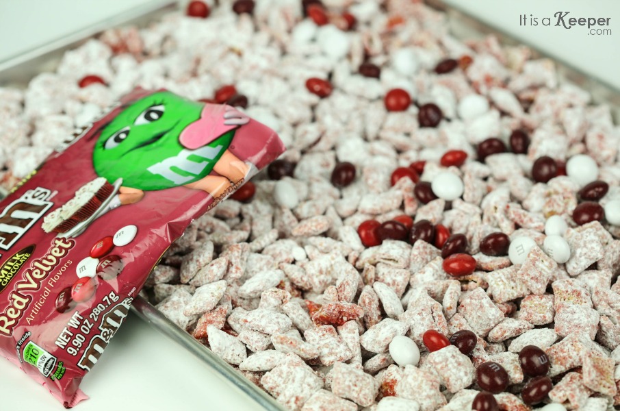 Red Velvet Muddy Buddies on a cookie sheet, with M&M being added to the baking sheet.