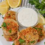 Plate of salmon patties garnished with parsley, served with a bowl of creamy dipping sauce. Reminiscent of tuna cakes, these delights are paired perfectly with lemon wedges and parsley sprigs in the background.