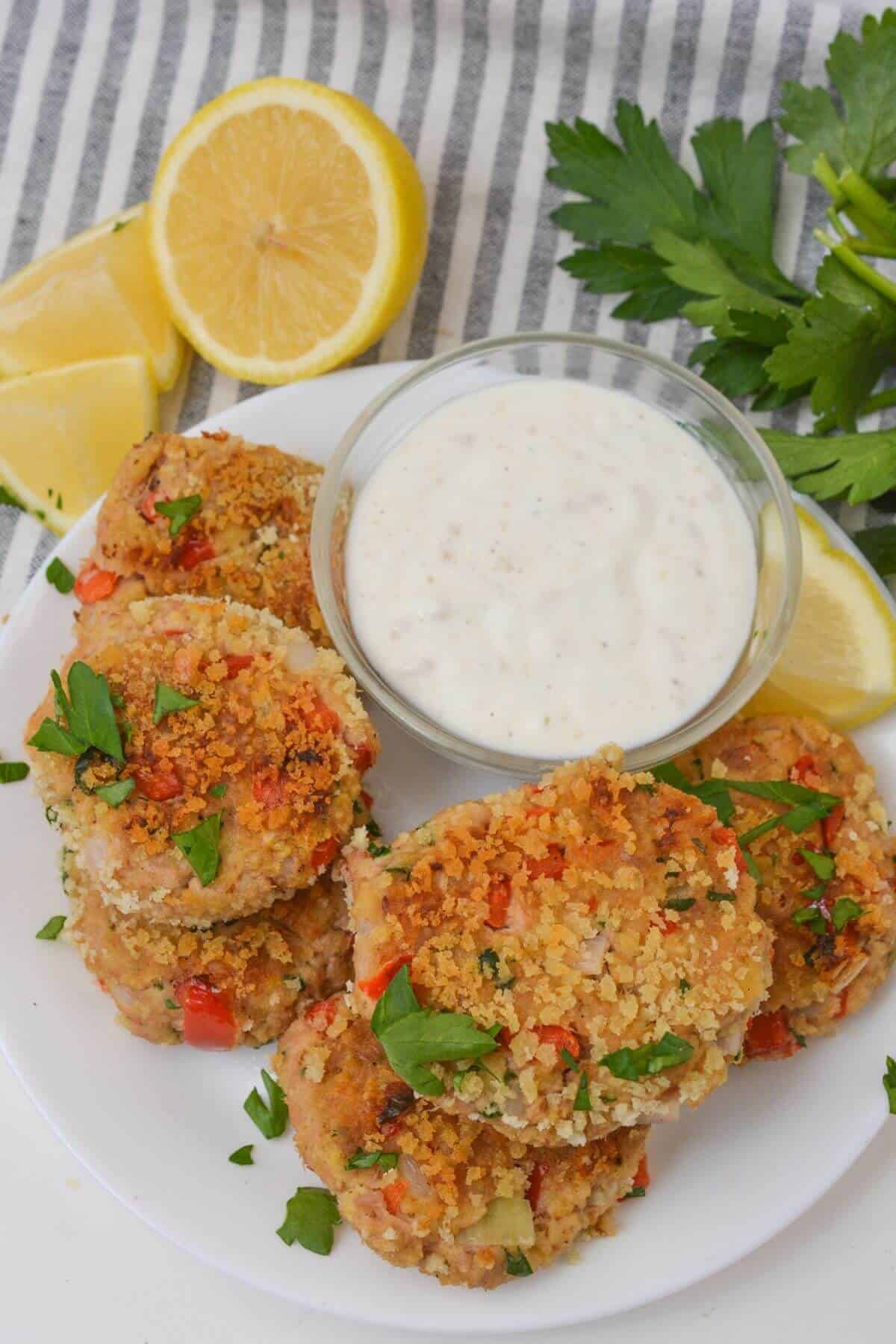 Plate of tuna patties garnished with parsley, served with a bowl of creamy dipping sauce. Reminiscent of tuna cakes, these delights are paired perfectly with lemon wedges and parsley sprigs in the background.