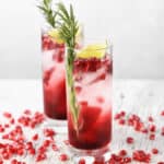 Two glasses of pomegranate cocktail with ice, garnished with rosemary and lime wedges, surrounded by pomegranate seeds on a wooden surface.