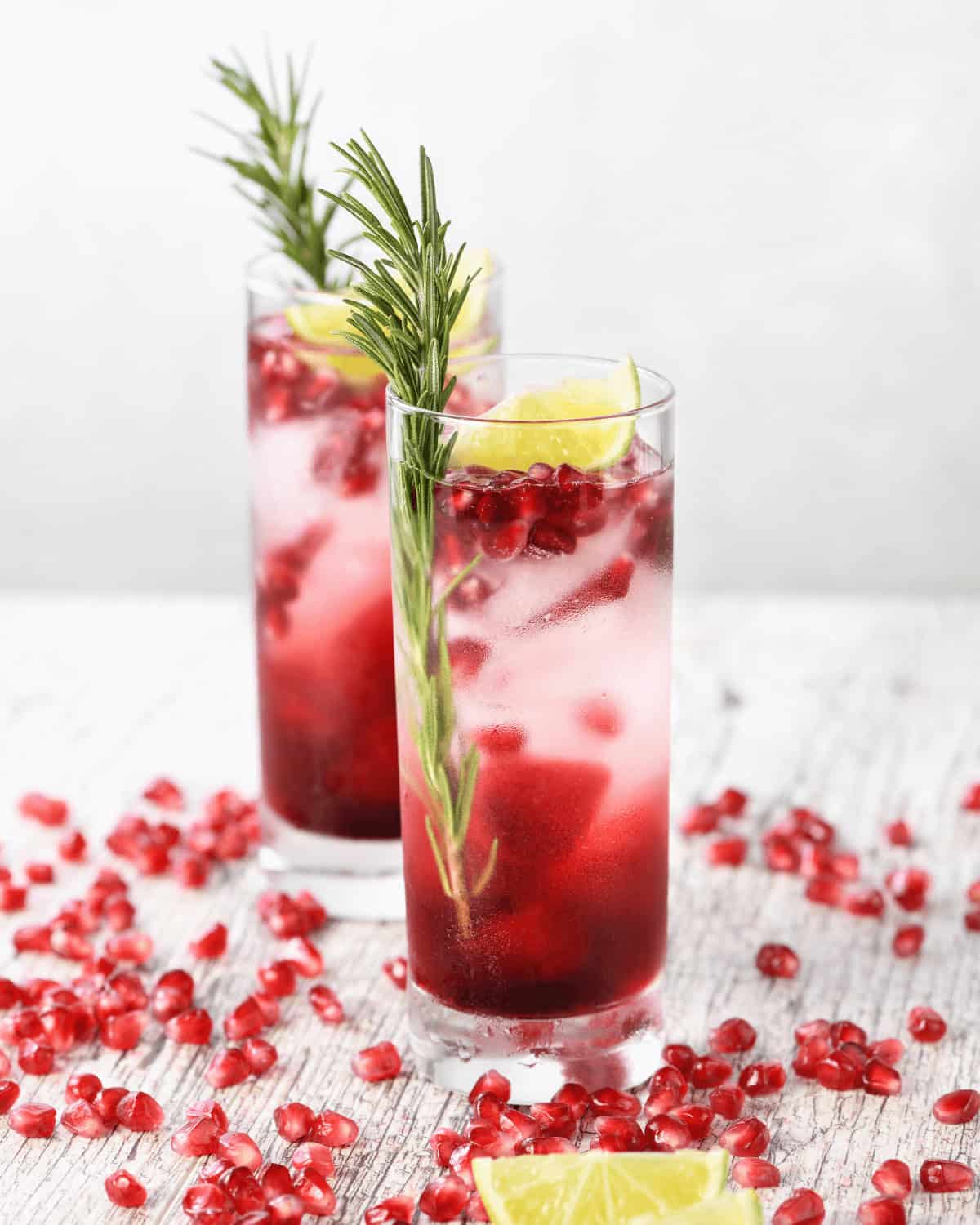 Two glasses of pomegranate cocktail with ice, garnished with rosemary and lime wedges, surrounded by pomegranate seeds on a wooden surface.