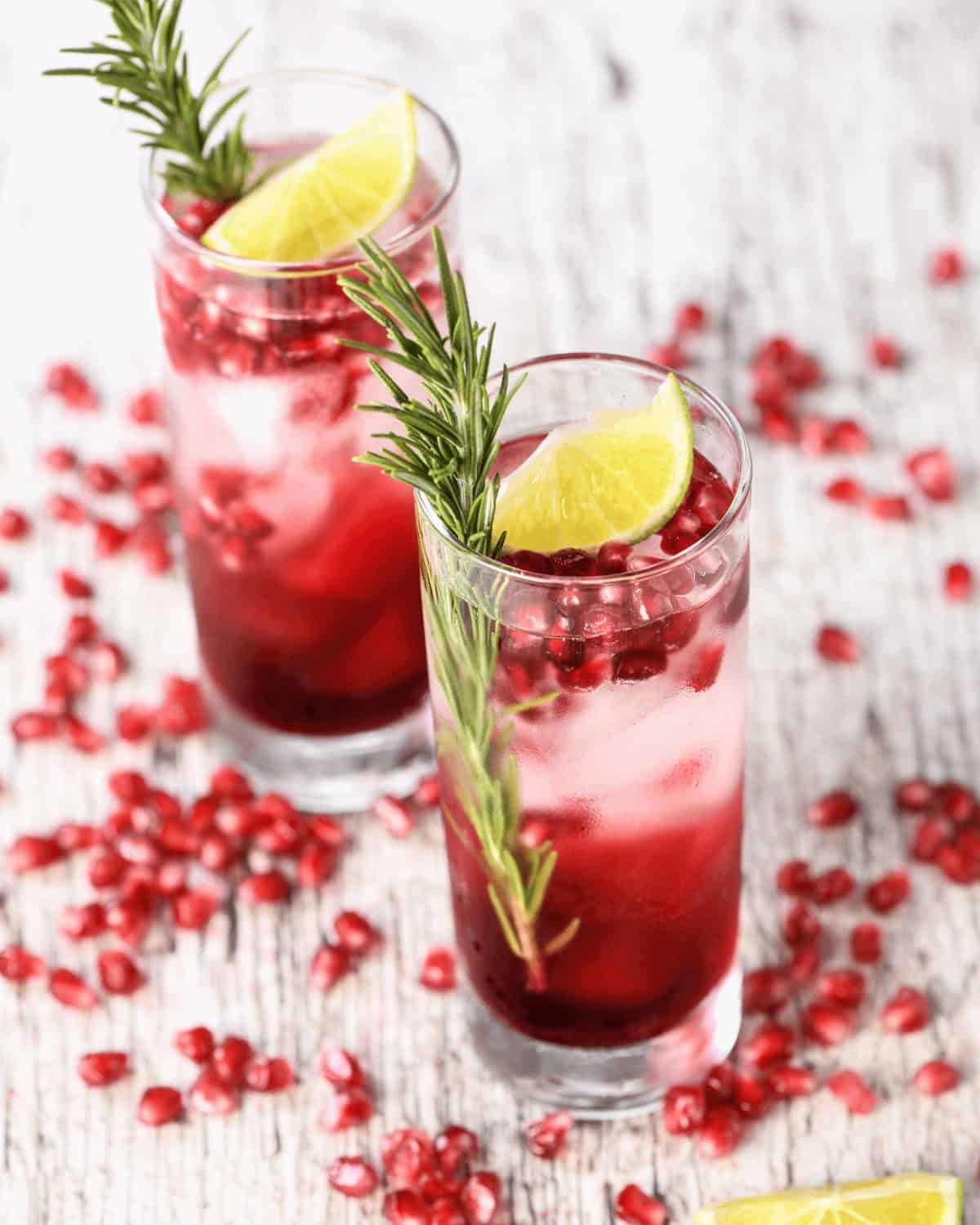 Two glasses of pomegranate cocktail with lime slices and rosemary sprigs on a rustic wooden surface, scattered with pomegranate seeds.