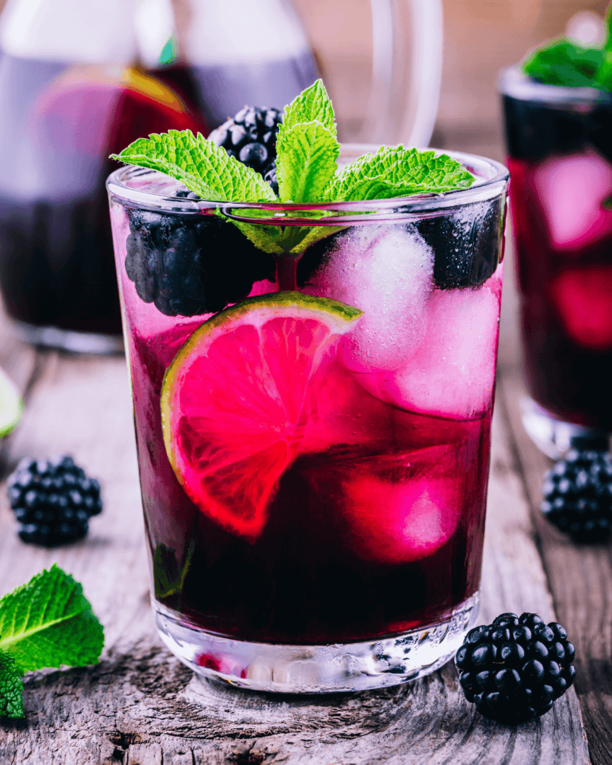 A glass of berry cocktail with ice, garnished with a lime slice, blackberries, and mint leaves on a wooden surface, reminiscent of a refreshing Blackberry Bourbon Smash.