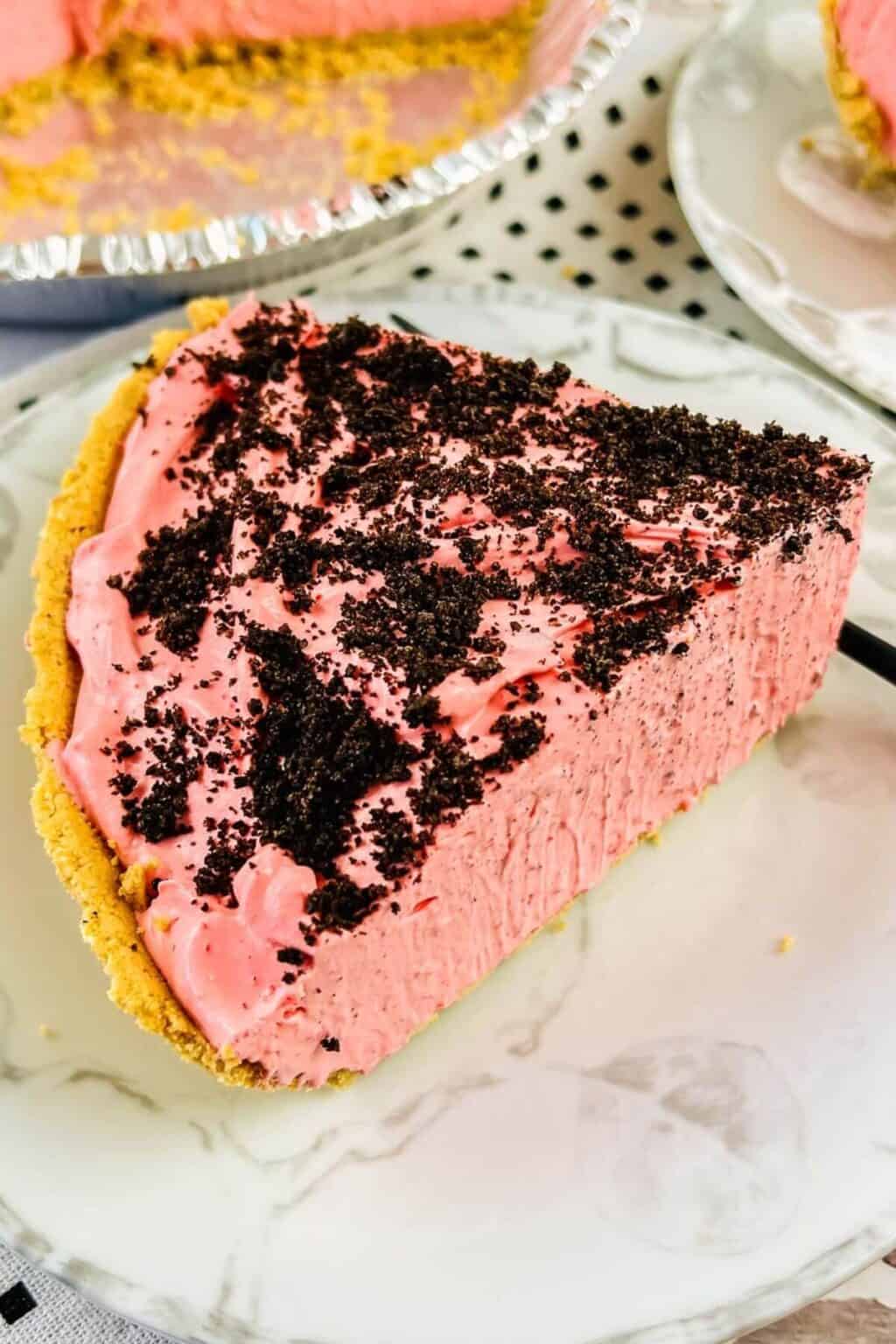 A slice of raspberry cream pie with a graham cracker crust and chocolate cookie crumbs on top, served on a white plate.
