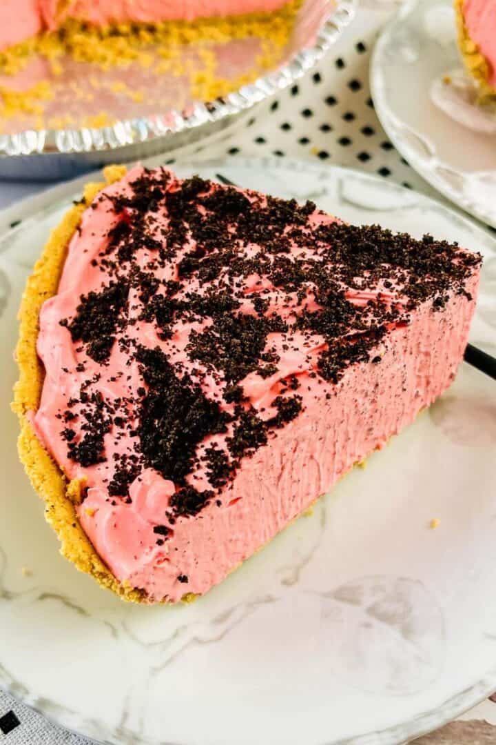 A slice of raspberry cream pie with a graham cracker crust and chocolate cookie crumbs on top, served on a white plate.