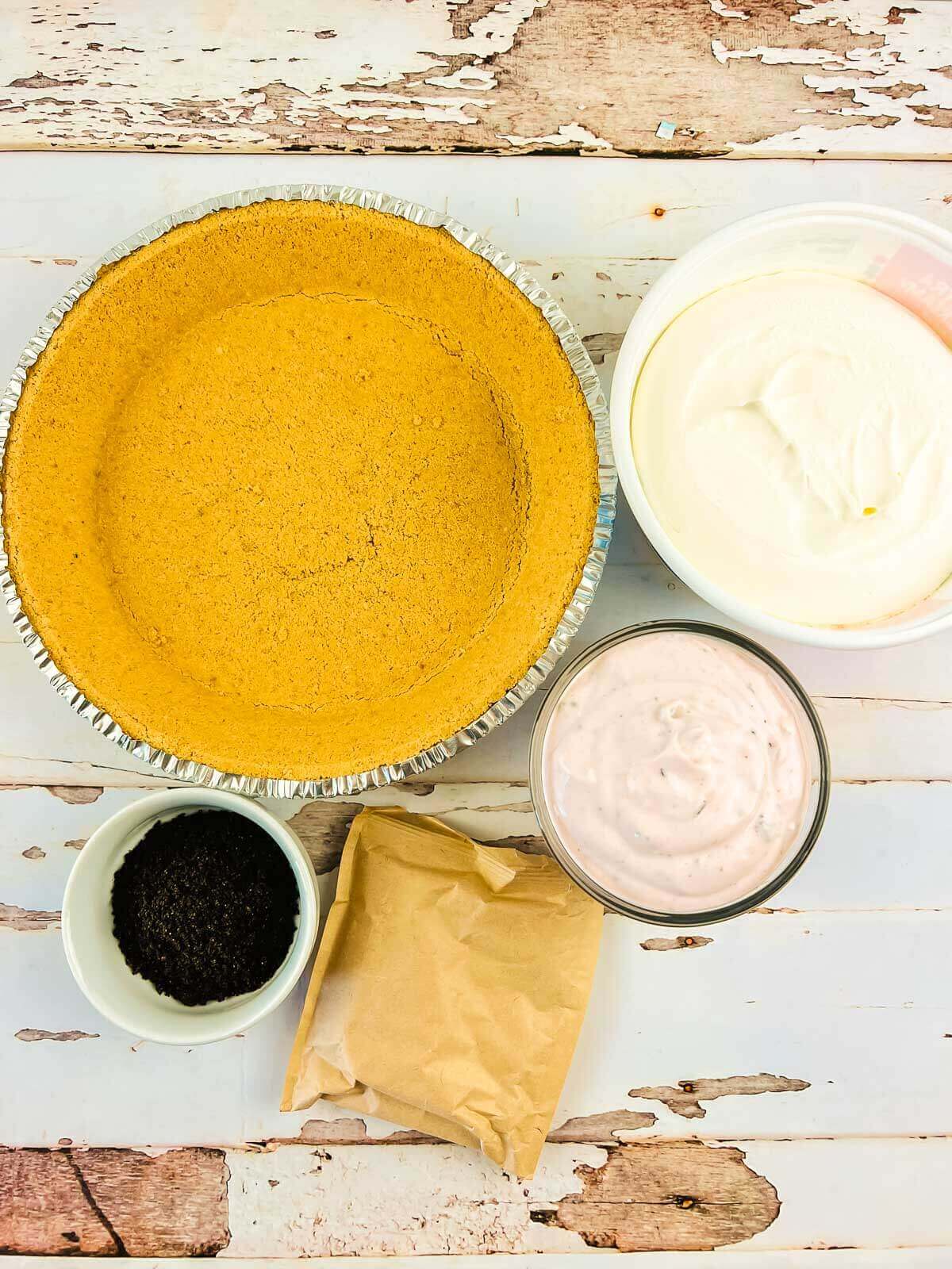 A graham cracker pie crust, a bowl of white creamy filling, a bowl of pink raspberry cream pie filling, a small bowl of crushed cookies, and a packet of powder on a rustic white surface.