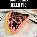 A slice of raspberry cream pie with a graham cracker crust and chocolate cookie crumbs on top sits on a white plate, accompanied by a black-handled fork.