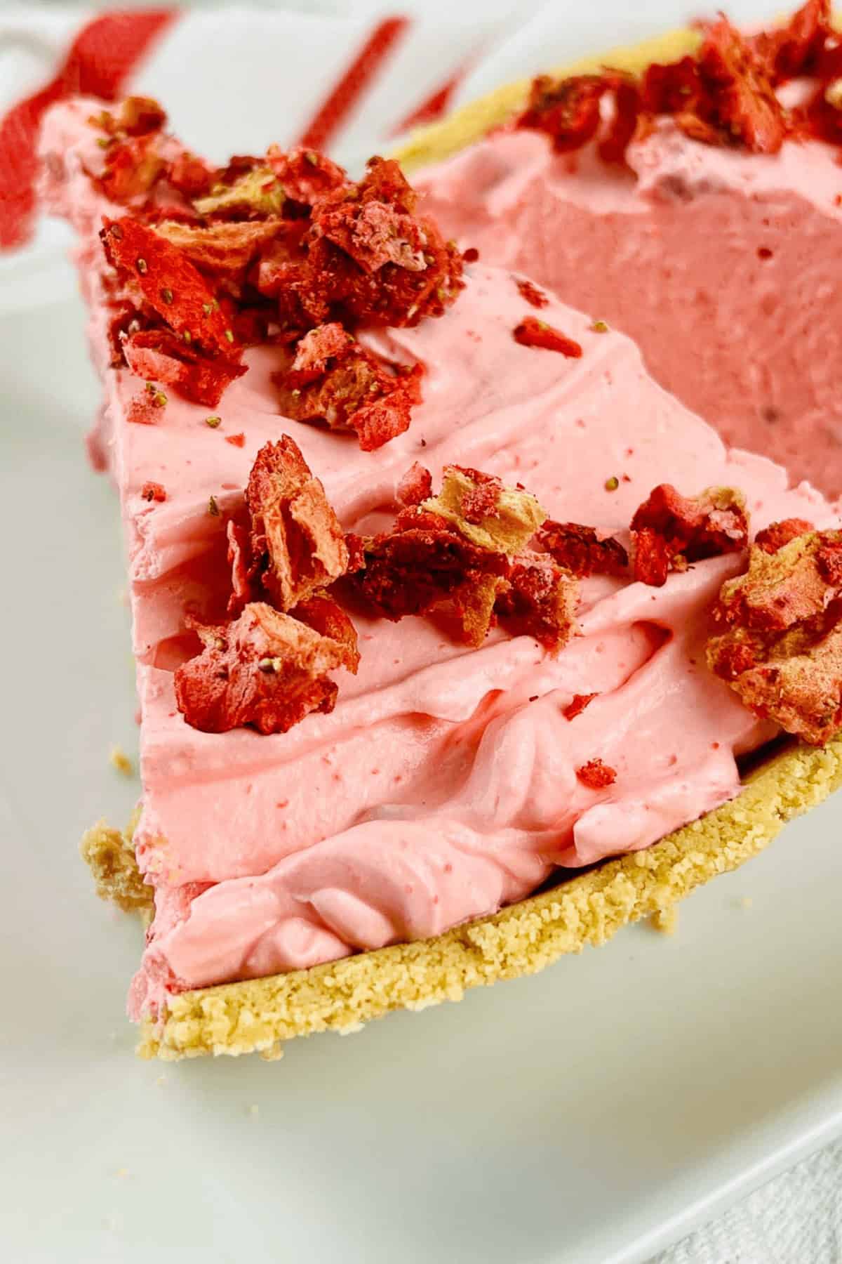 A slice of strawberry pie with graham cracker crust, topped with red crumbled pieces and set in a crumbly crust on a white plate.