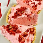 Three slices of dessert topped with crushed strawberries are arranged on a rectangular white platter with a red and white striped cloth underneath.