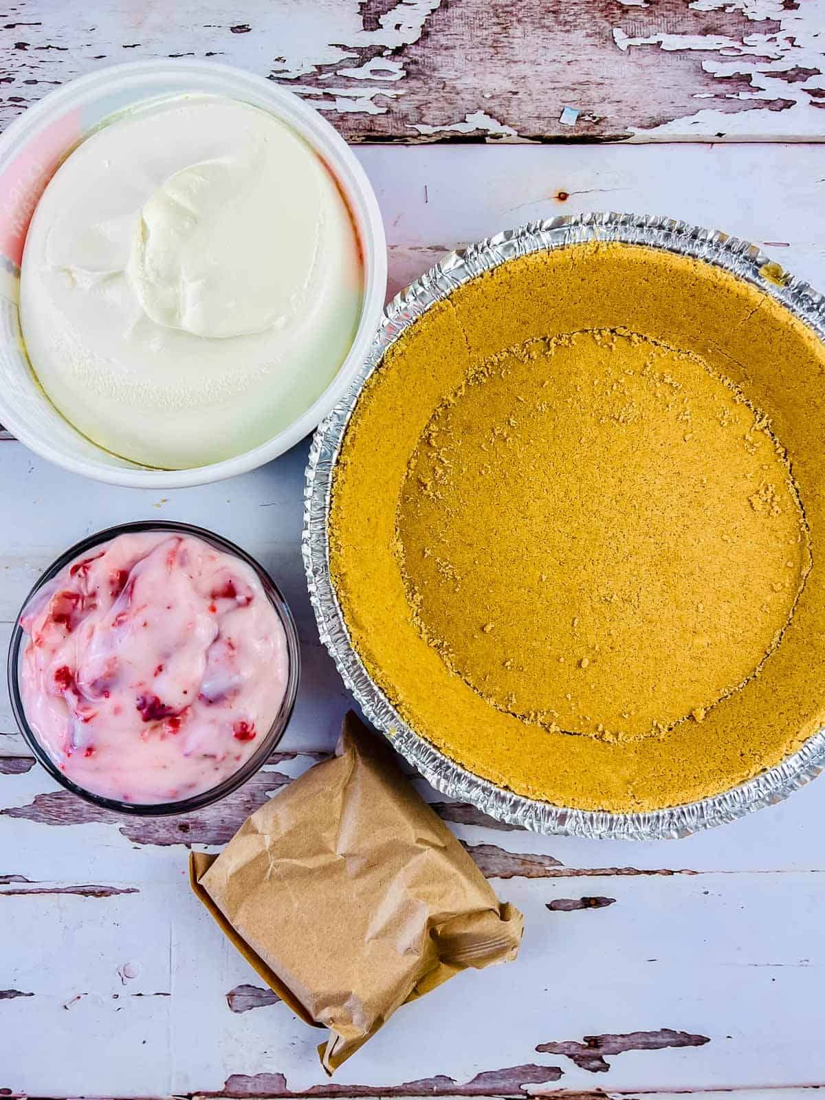 An unfilled graham cracker pie crust next to bowls of whipped cream and a pink fruit mixture, ready to become a delicious strawberry pie with graham cracker crust. A small brown paper bag completes the scene on a rustic, chipped wooden surface.