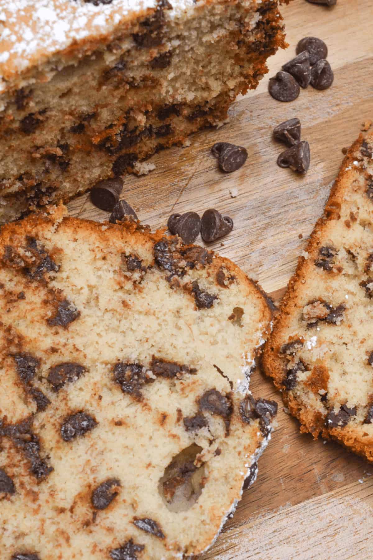 Two slices of chocolate chip banana bread rest on a wooden surface, their rich aroma mingling with scattered chocolate chips.
