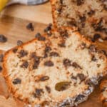 A slice of banana chocolate chip bread rests on a wooden surface, with a whole loaf and scattered chocolate chips adorning the background.