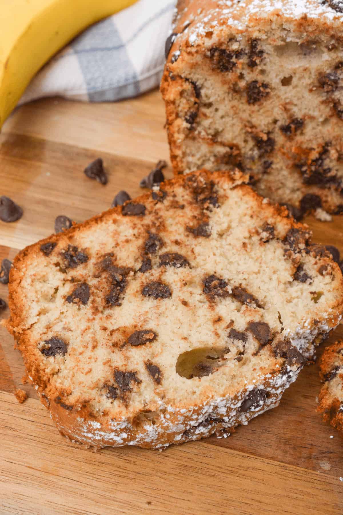 A slice of banana chocolate chip bread rests on a wooden surface, with a whole loaf and scattered chocolate chips adorning the background.