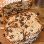A sliced loaf of chocolate chip banana bread rests on a wooden board, lightly dusted with powdered sugar, with a metal shaker nearby.