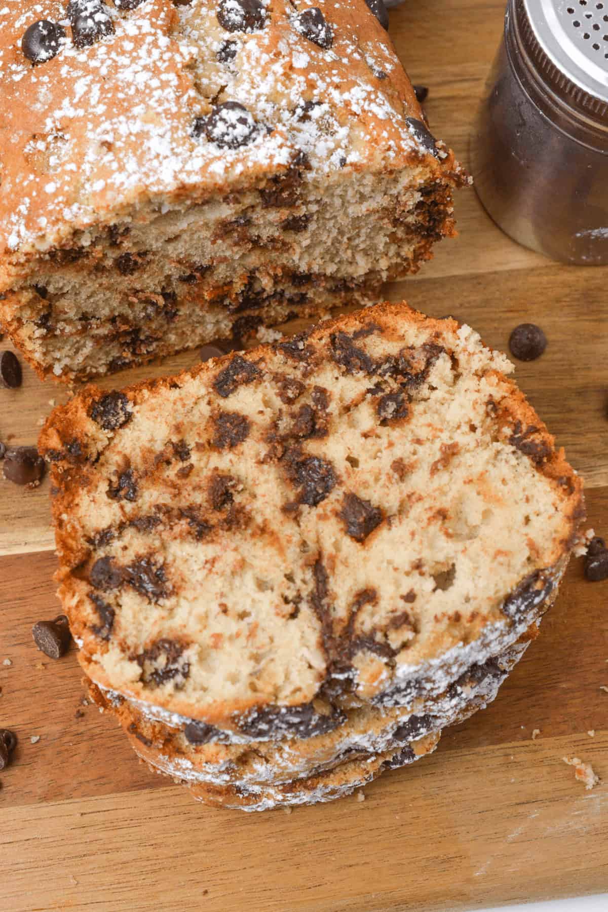 A sliced loaf of chocolate chip banana bread rests on a wooden board, lightly dusted with powdered sugar, with a metal shaker nearby.