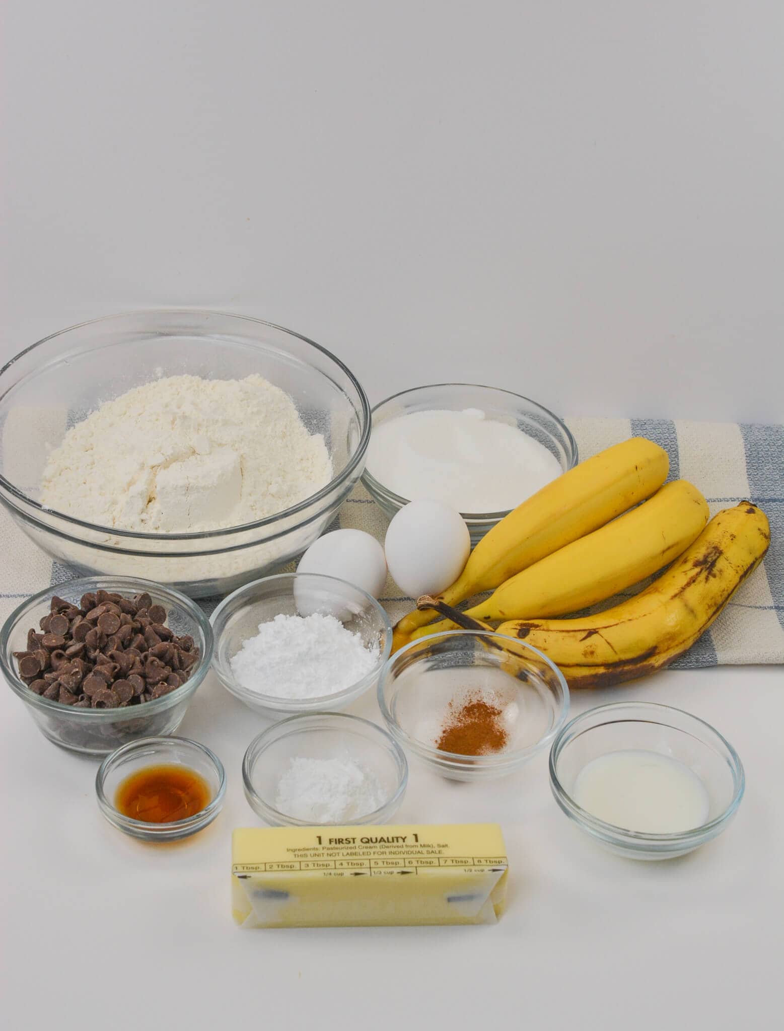 Ingredients of flour, sugar, eggs, ripe bananas, chocolate chips, butter, vanilla extract, baking powder, salt, cinnamon, and milk are arranged neatly on a counter with a striped cloth.