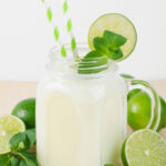 A refreshing lime and coconut mojito smoothie in a mason jar with a white and green striped straw, garnished with lime slices and mint leaves, surrounded by whole and halved limes