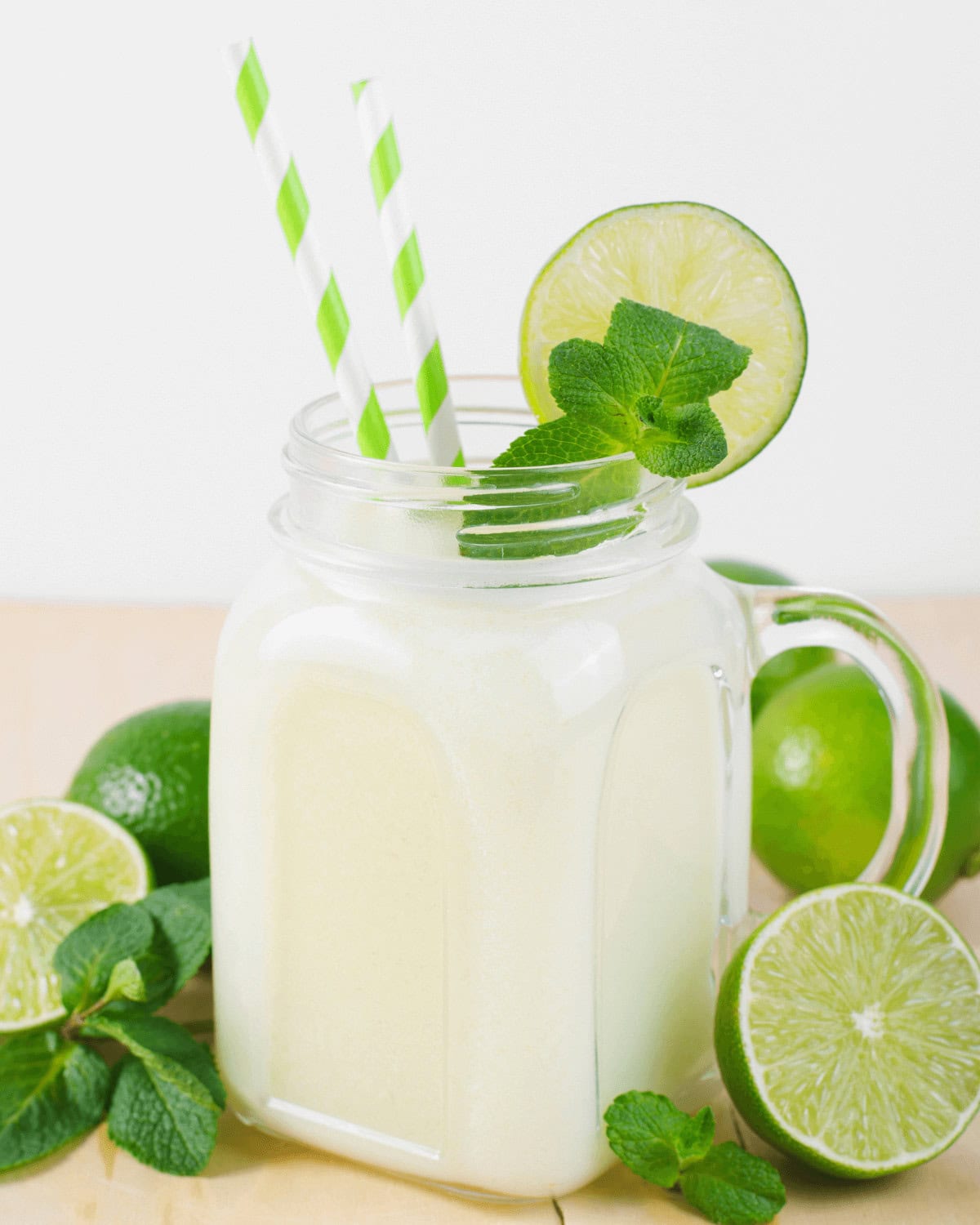A refreshing lime and coconut mojito smoothie in a mason jar with a white and green striped straw, garnished with lime slices and mint leaves, surrounded by whole and halved limes
