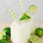 Two jars of lime coconut mojito milkshake with striped straws, garnished with lime slices and mint, surrounded by whole limes on a light background.