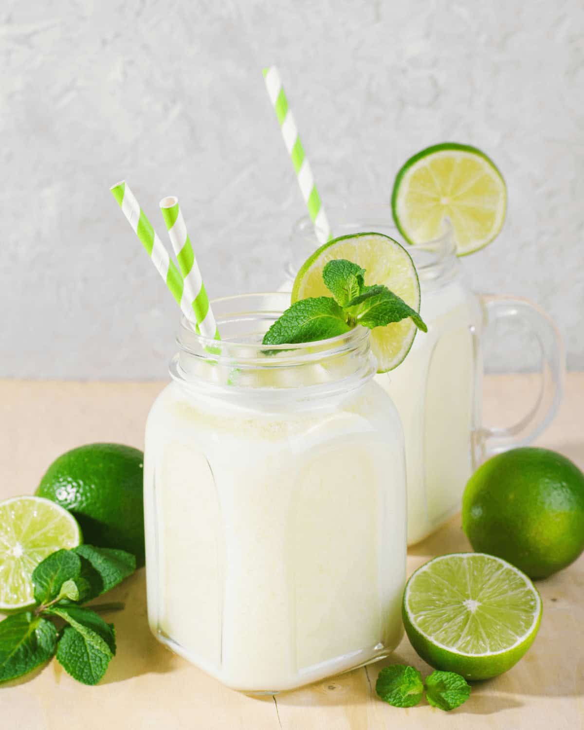 Two jars of lime coconut mojito milkshake with striped straws, garnished with lime slices and mint, surrounded by whole limes on a light background.