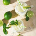 Two glasses with straws, garnished with lime slices and mint, surrounded by fresh limes on a wooden surface.