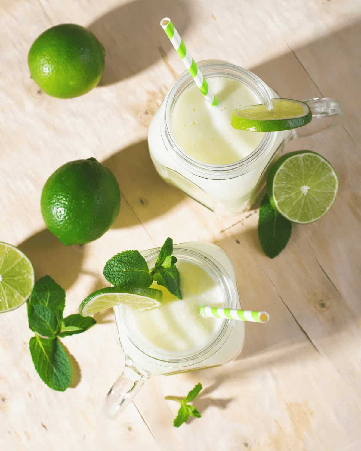 Two glasses with straws, garnished with lime slices and mint, surrounded by fresh limes on a wooden surface.