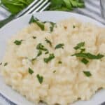 A luscious plate of creamy risotto, garnished with chopped parsley, rests on a striped cloth beside a fork and fresh parsley sprigs.