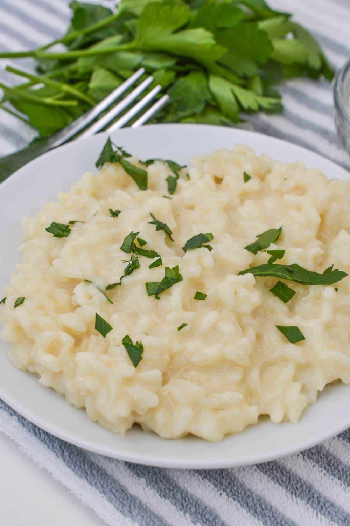 A luscious plate of creamy risotto, garnished with chopped parsley, rests on a striped cloth beside a fork and fresh parsley sprigs.