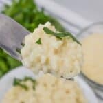 A fork holding a bite of luscious side perfectly creamy and garnished with chopped herbs, hovers before a blurred background showcasing a full plate, a bowl of grated cheese, and vibrant green leaves.