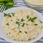 A plate of perfectly prepared creamy risotto garnished with chopped parsley sits on a striped cloth, accompanied by a bowl of grated cheese and fresh parsley sprigs.
