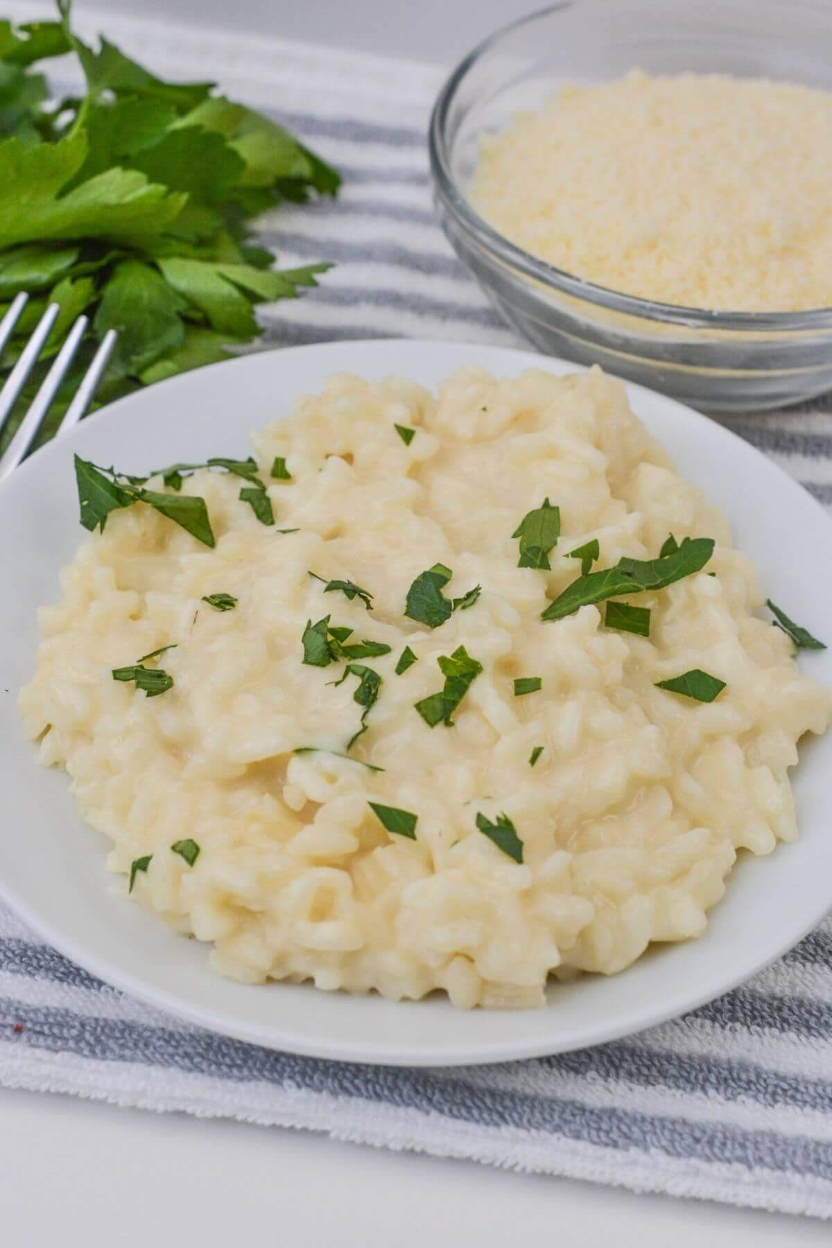 A plate of perfectly prepared creamy risotto garnished with chopped parsley sits on a striped cloth, accompanied by a bowl of grated cheese and fresh parsley sprigs.